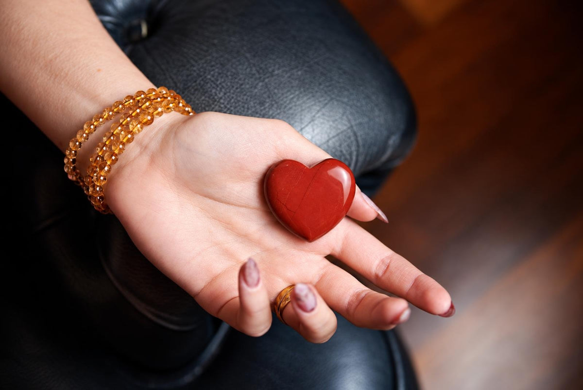 A polished red heart-shaped stone rests in an open hand wearing an orange beaded bracelet.