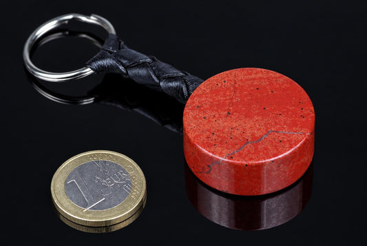 A red cylindrical stone with fine dark markings is attached to a black braided strap and metal keyring beside a coin on a reflective black surface.