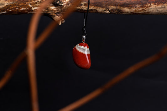 A polished red jasper pendant with a white band hangs from a black cord on a wooden branch against a dark background.