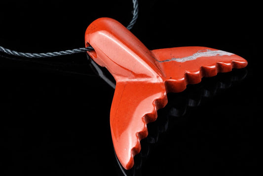 A polished red jasper pendant shaped like a dolphin tail, with a silver-toned vein and black cord on a dark surface.