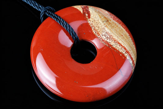 A polished red jasper pendant with a central hole, featuring cream and brown banding on one side, threaded with a dark blue cord against a black background.