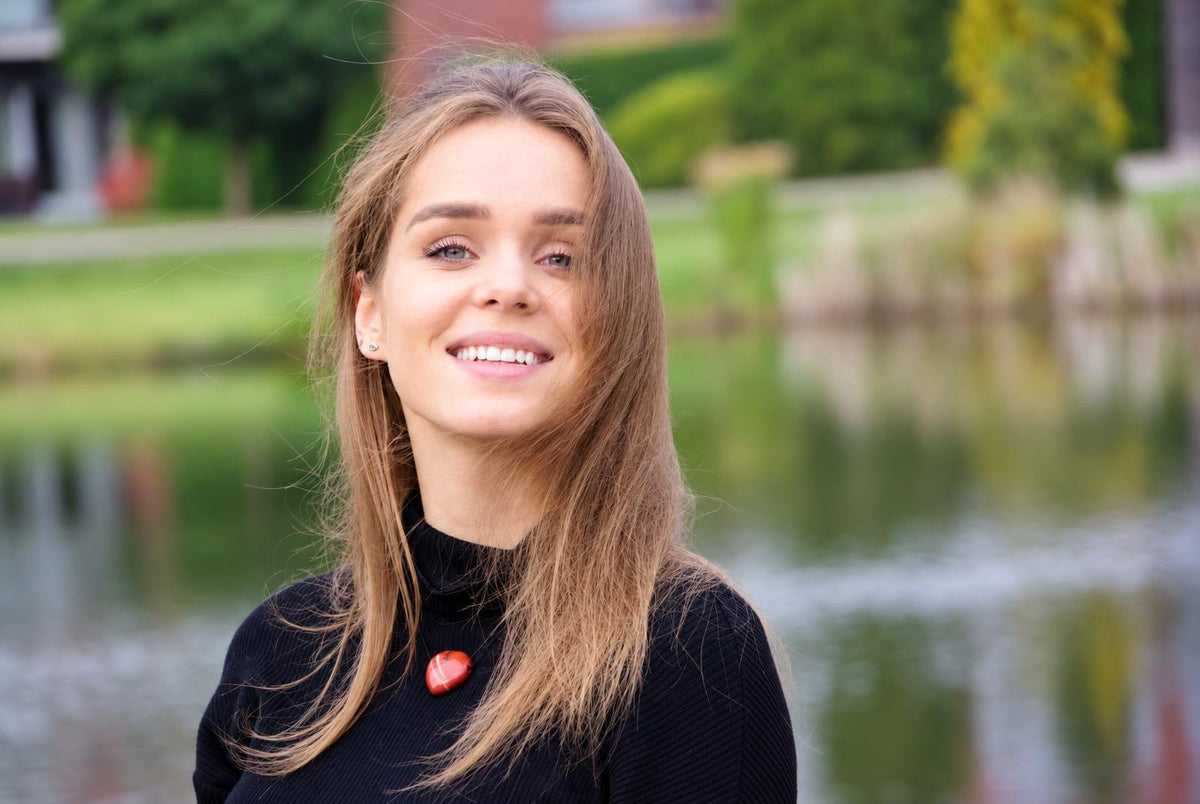 A smiling woman with long light brown hair wears a black turtleneck and a red oval pendant on a dark surface.