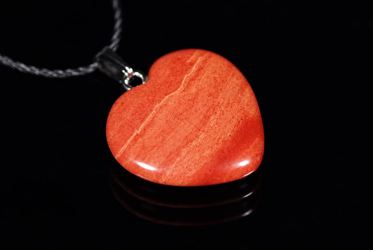 A polished red jasper heart pendant with a silver-toned bail and chain, resting on a dark reflective surface.
