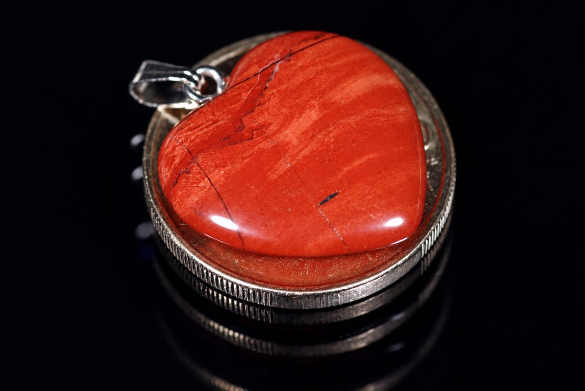 A silver-toned pendant with a polished, heart-shaped red jasper stone set in a circular bezel on a dark reflective surface.