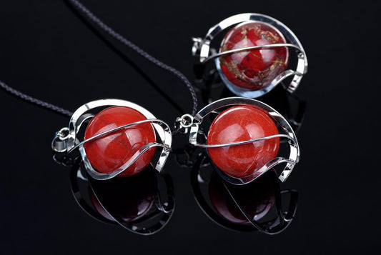 Three red jasper sphere pendants with silver-toned wire frames on dark cords, arranged on a black reflective surface.