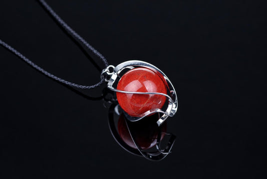 A silver-toned wire pendant holds a polished red sphere on a dark cord, resting on a reflective black surface.