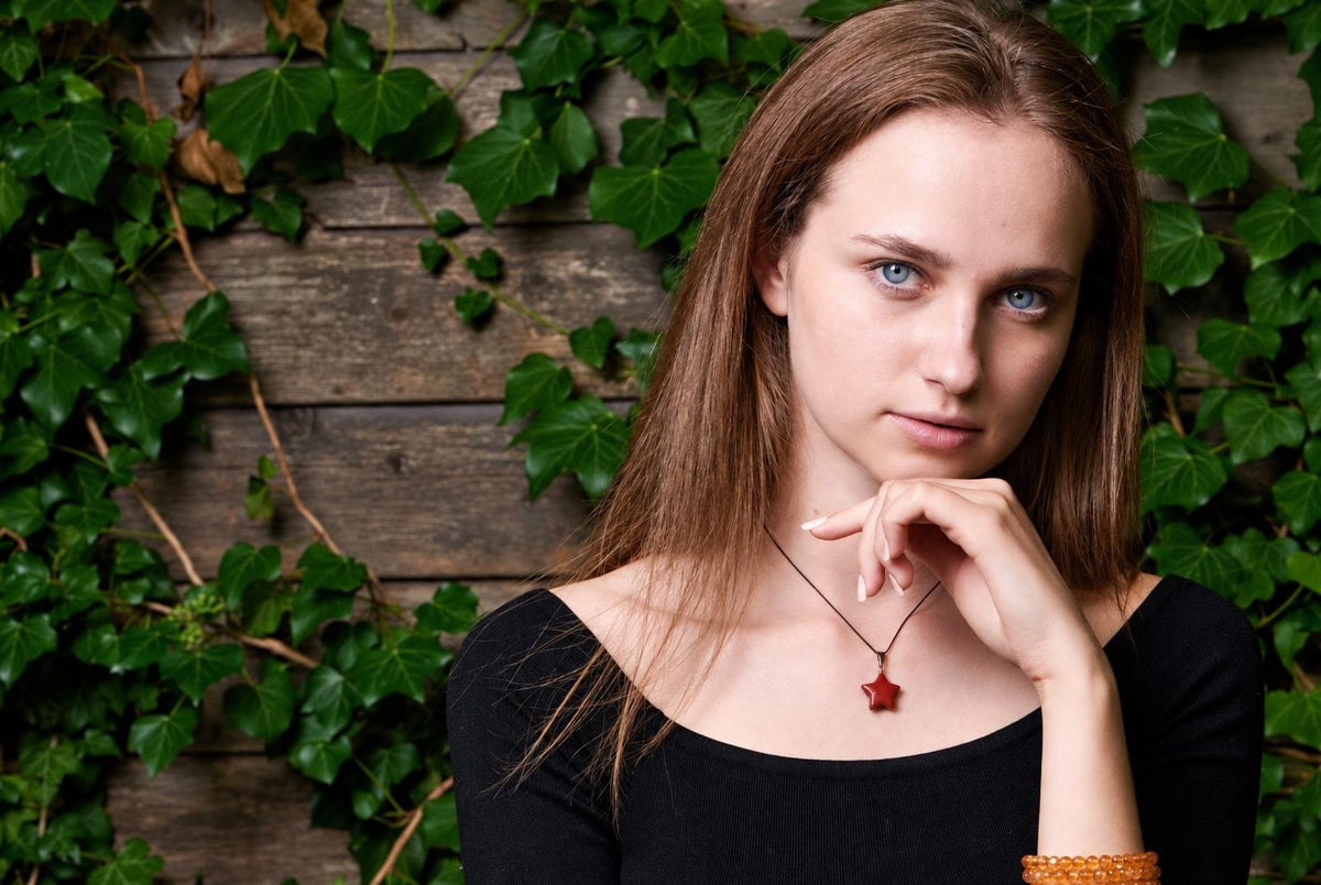 A woman wears a black top and a necklace with a red star pendant against a wooden wall with green ivy.