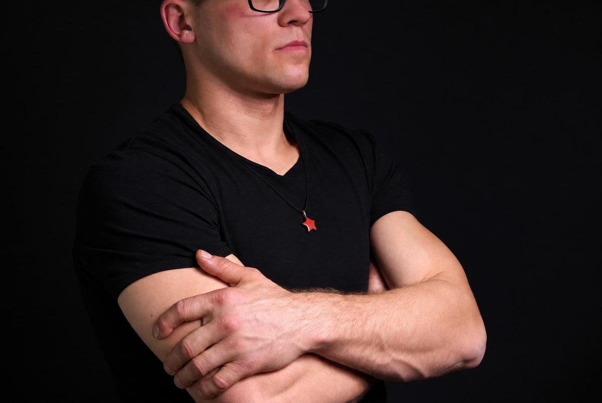 A man in a black t-shirt wears a necklace with a small red star pendant against a dark background.