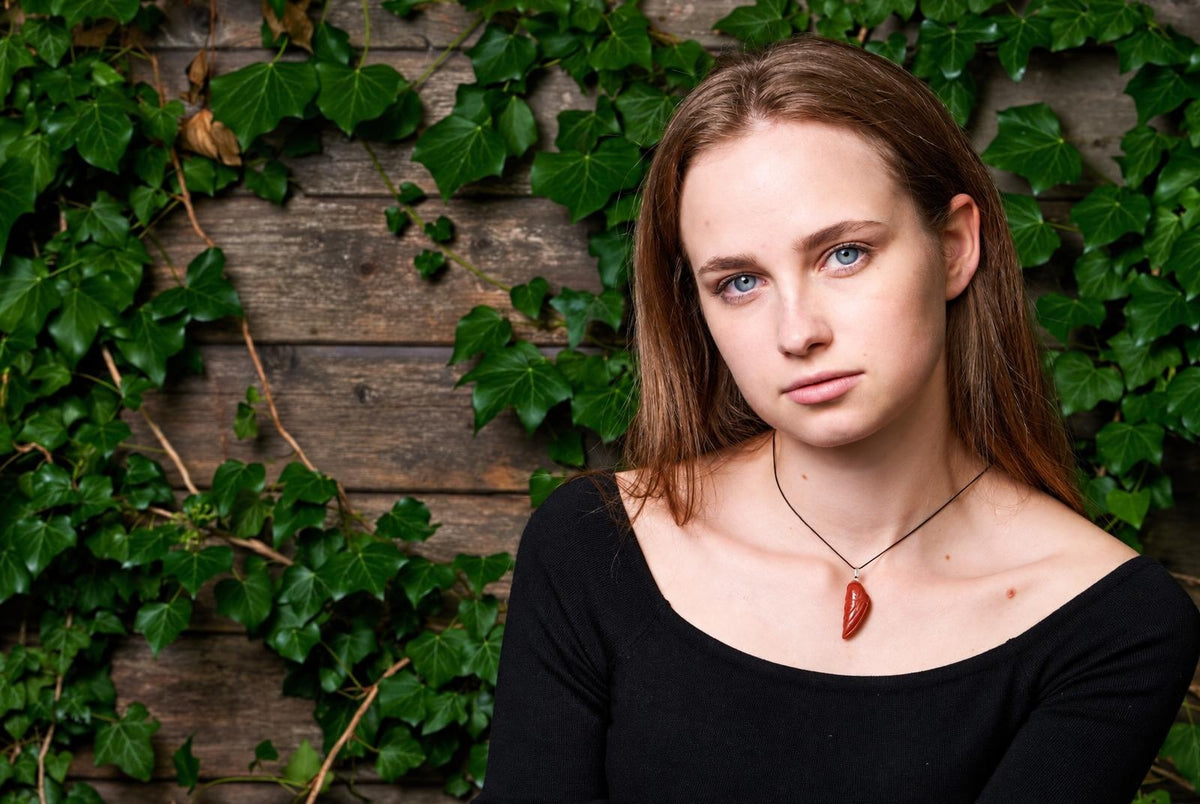 A woman wears a red jasper pendant shaped like a wing on a black cord, against a wooden wall with green ivy.