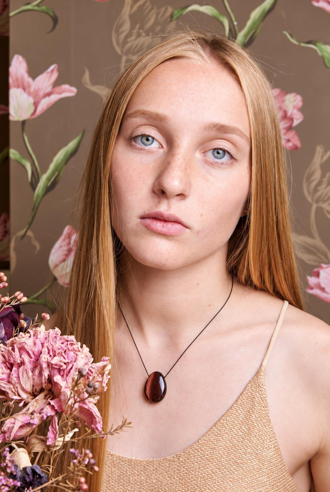 A woman wears a necklace with a smooth, dark reddish-brown oval pendant on a thin black cord against a floral wallpaper background.