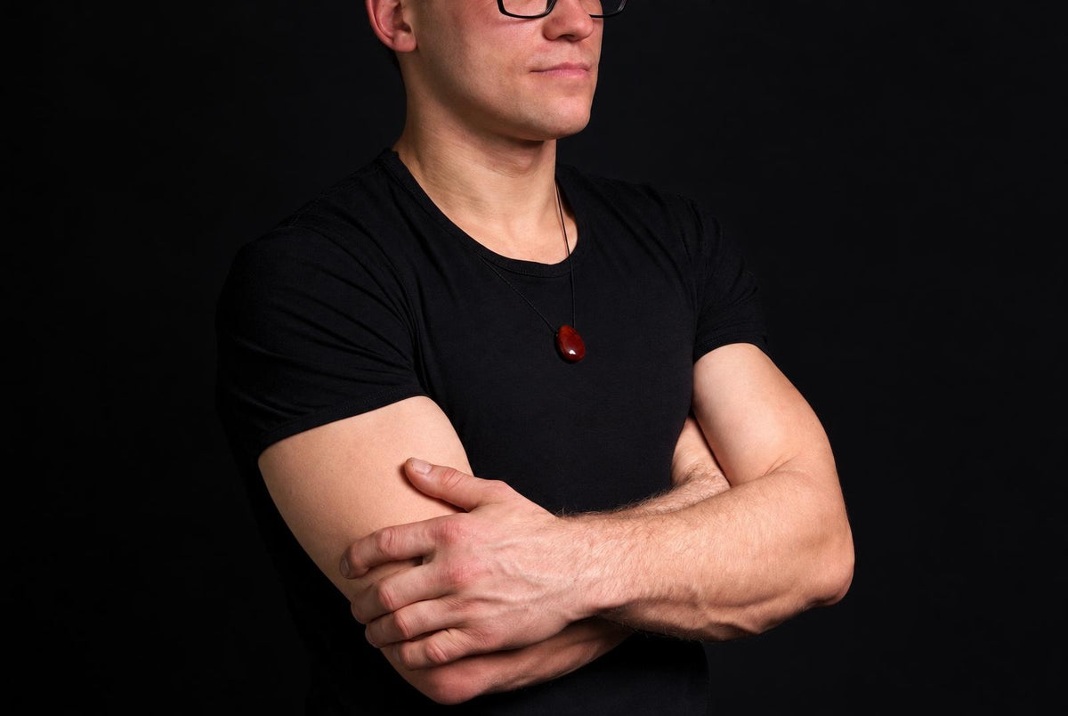 A man in a black t-shirt wears a necklace with a smooth, oval red pendant against a dark background.