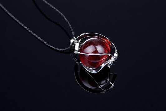 A silver-toned pendant with a round, red sphere on a dark cord against a black reflective surface.