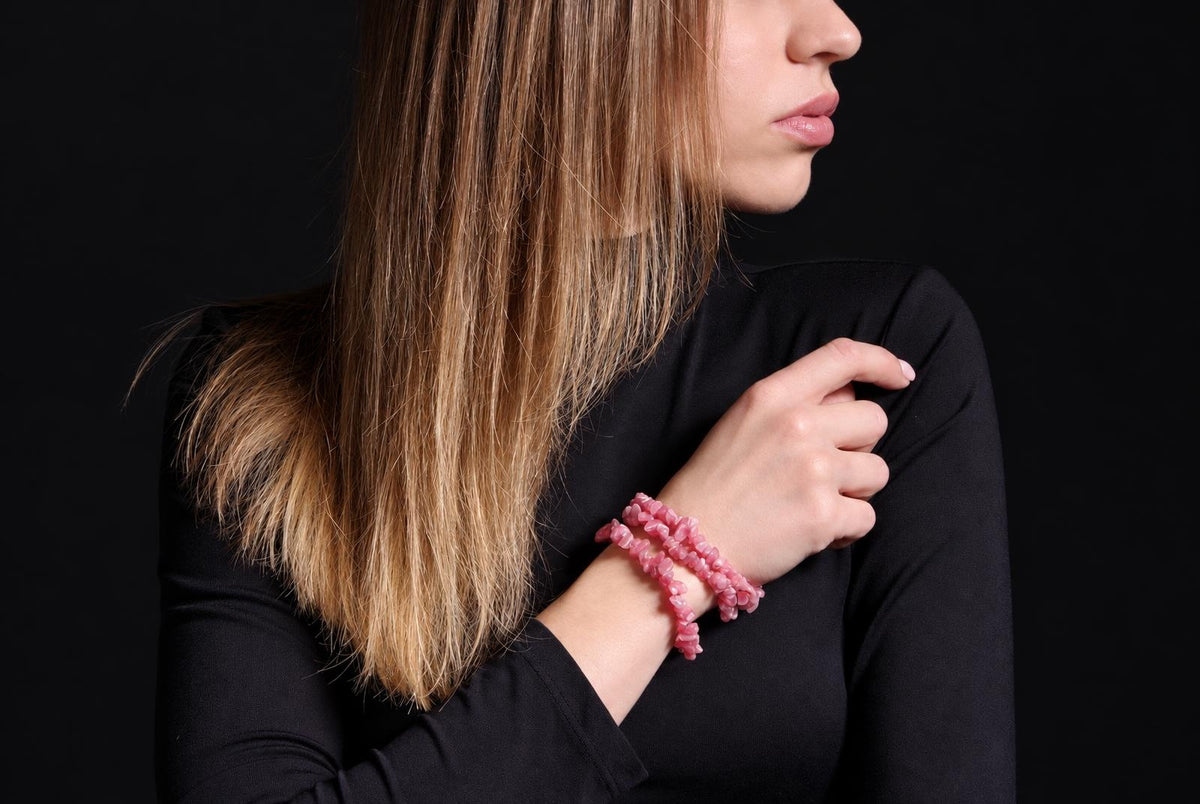 A woman with blonde hair wears a pink beaded bracelet on her wrist against a dark background.