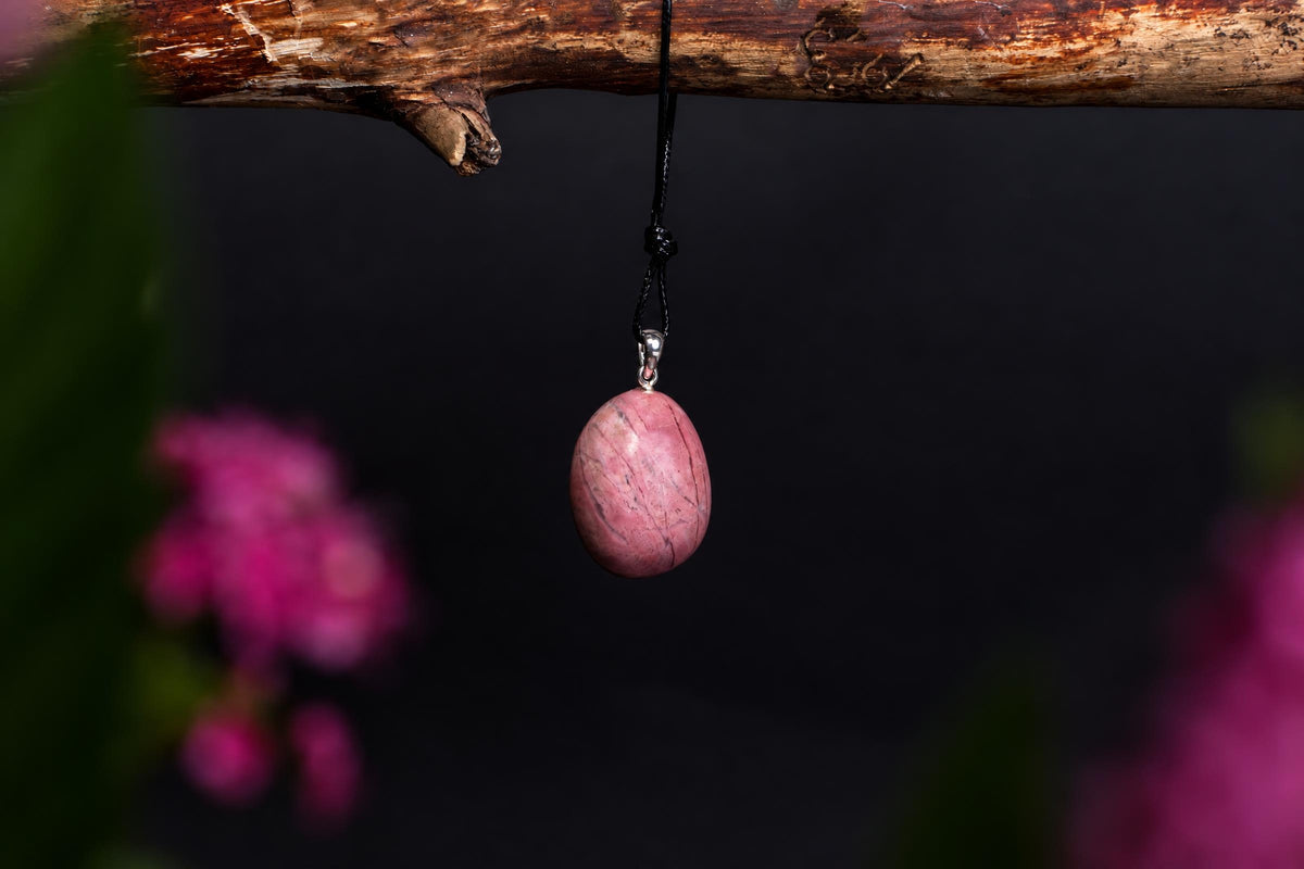 A pink, oval Rhodonite pendant with silver-toned bail hangs from a black cord on a wooden branch against a dark background.