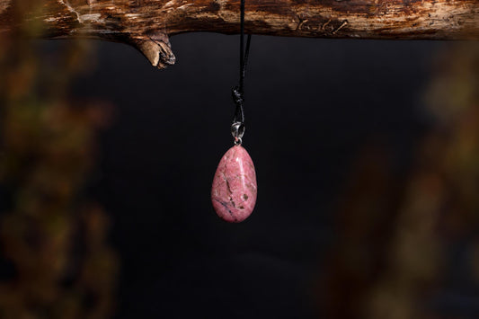 A pink rhodonite pendant with a silver bail hangs from a black cord on a wooden branch against a dark background.