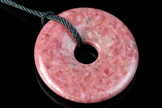 A pink, donut-shaped Rhodonite pendant with a central hole and a dark cord looped through it, set against a black background.