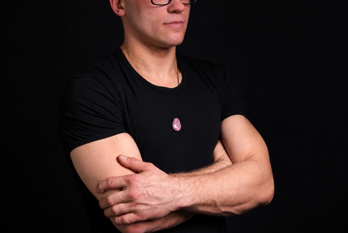 A man in a black t-shirt wears a necklace with a pinkish, oval Rhodonite pendant on a dark cord against a black background.