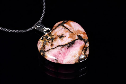 A heart-shaped Rhodonite pendant with pink, brown and black veining hangs from a silver-toned chain on a dark surface.