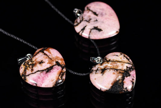 Three heart-shaped Rhodonite pendants with pink, brown and black veining on dark cords against a black reflective surface.