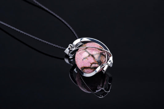 A silver-toned pendant with a pink and black patterned sphere on a dark cord, resting on a dark surface.