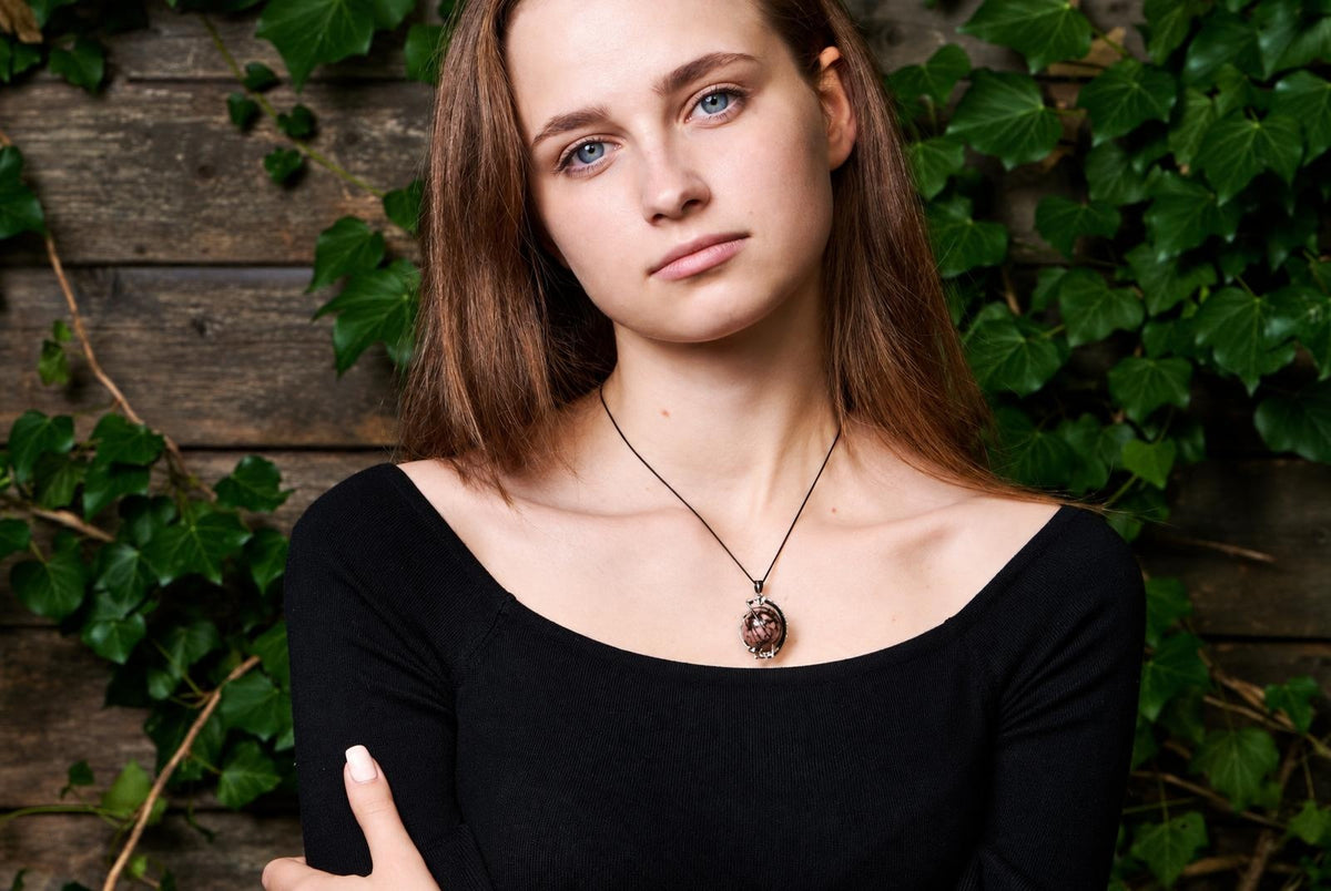 A woman wears a black top and a pendant with a round, brownish stone on a dark cord against a wooden wall with green ivy.