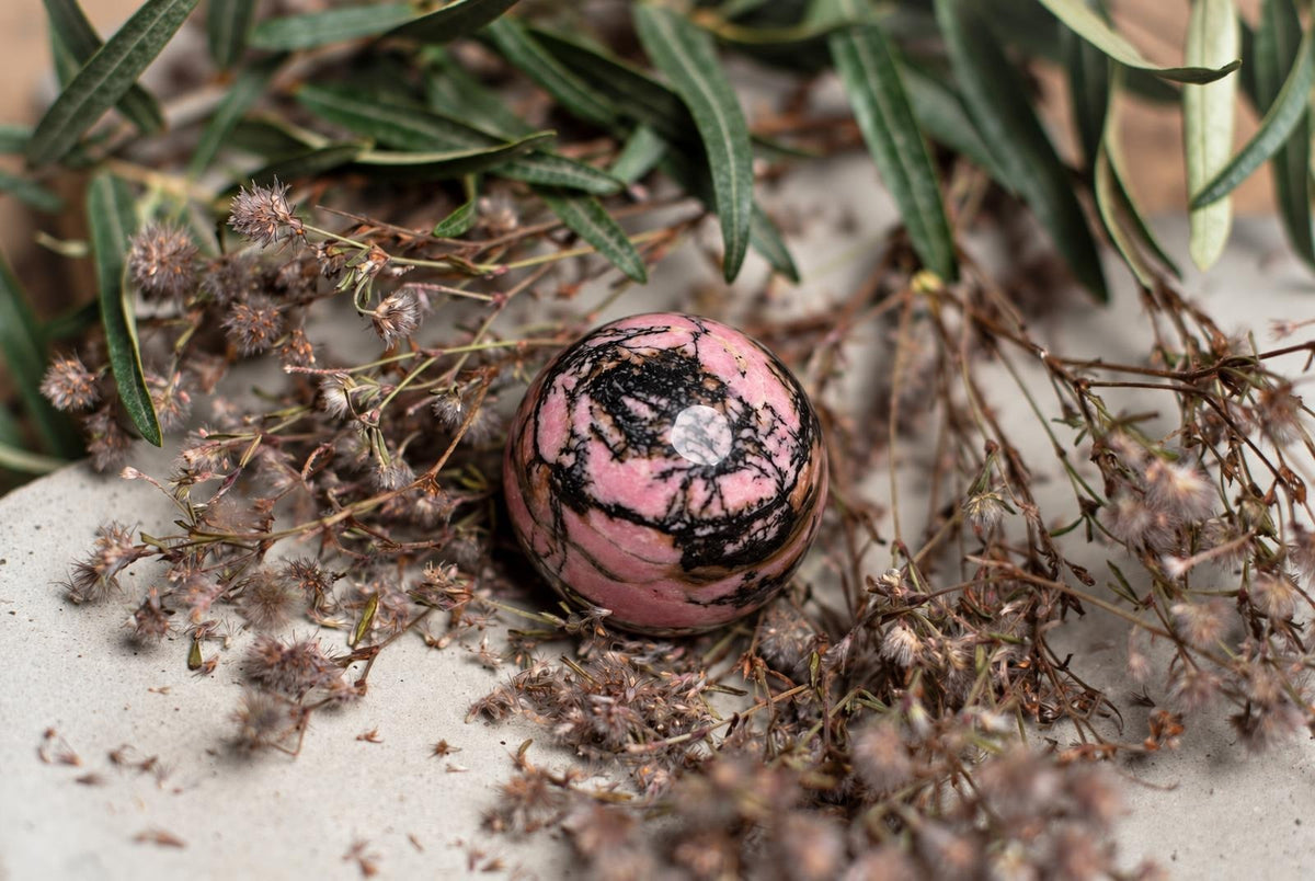 A polished Rhodonite sphere with pink and black swirls rests on a pale surface among green leaves and dried flowers.