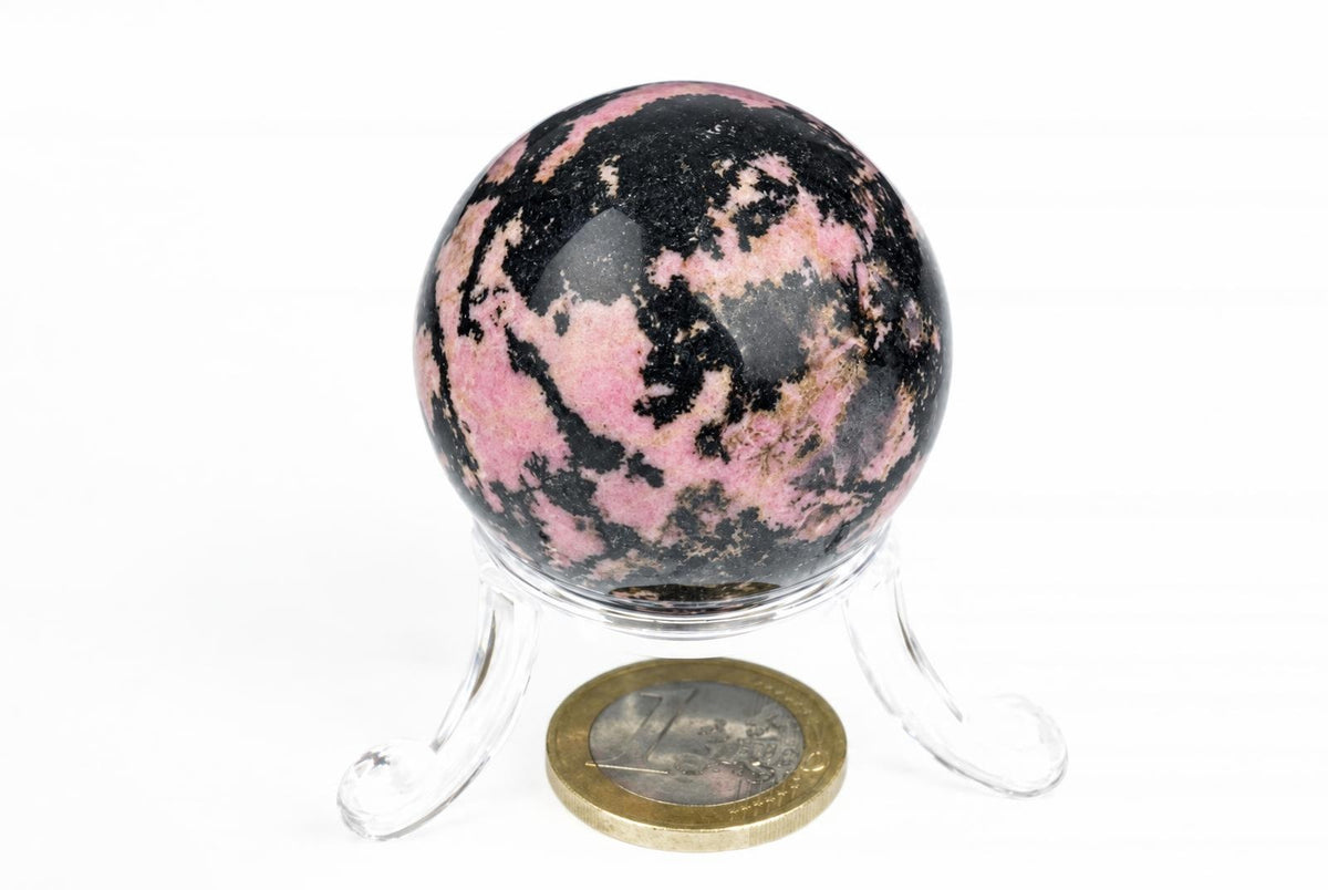 A polished Rhodonite sphere with pink and black patterns sits on a clear stand against a white background.