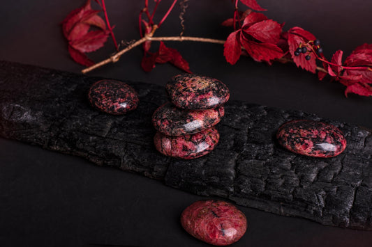 Five polished red and black speckled stones rest on a dark wooden surface with red leaves in the background.