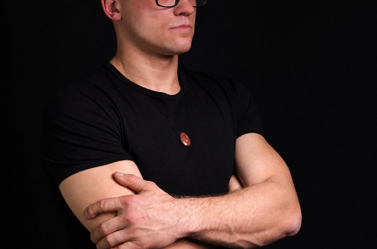 A man in a black t-shirt wears a pendant with an oval, reddish-brown stone on a dark surface.
