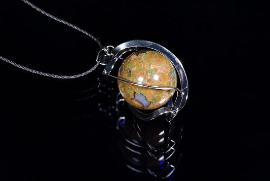 A silver-toned pendant with a twisted wire frame holds a polished, multi-coloured sphere of orange, green and blue stone on a dark reflective surface.
