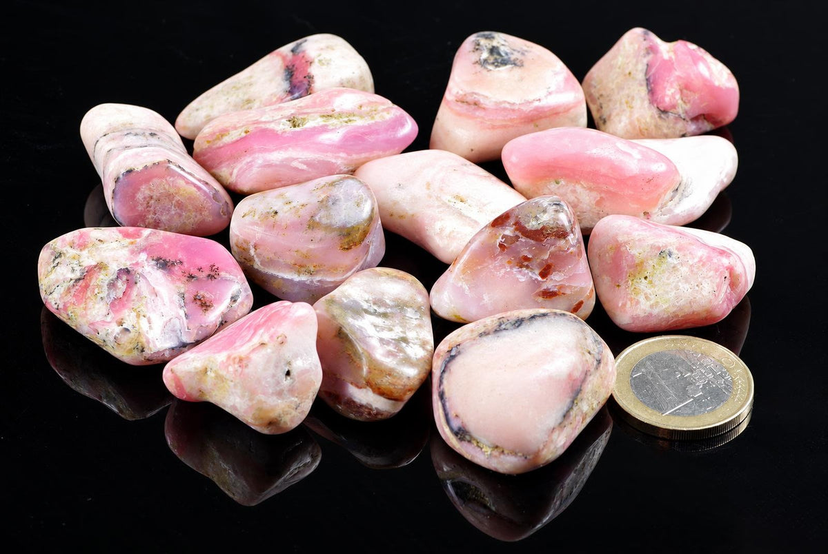 A collection of polished rose opal stones with pink, white and dark inclusions on a black reflective surface next to a coin.