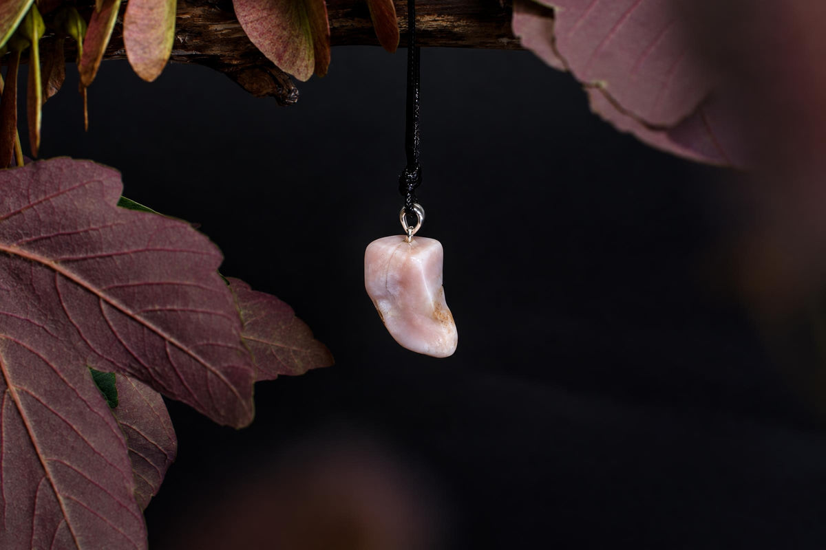 A pale pink, irregularly shaped stone pendant hangs from a black cord against a dark background with reddish leaves.