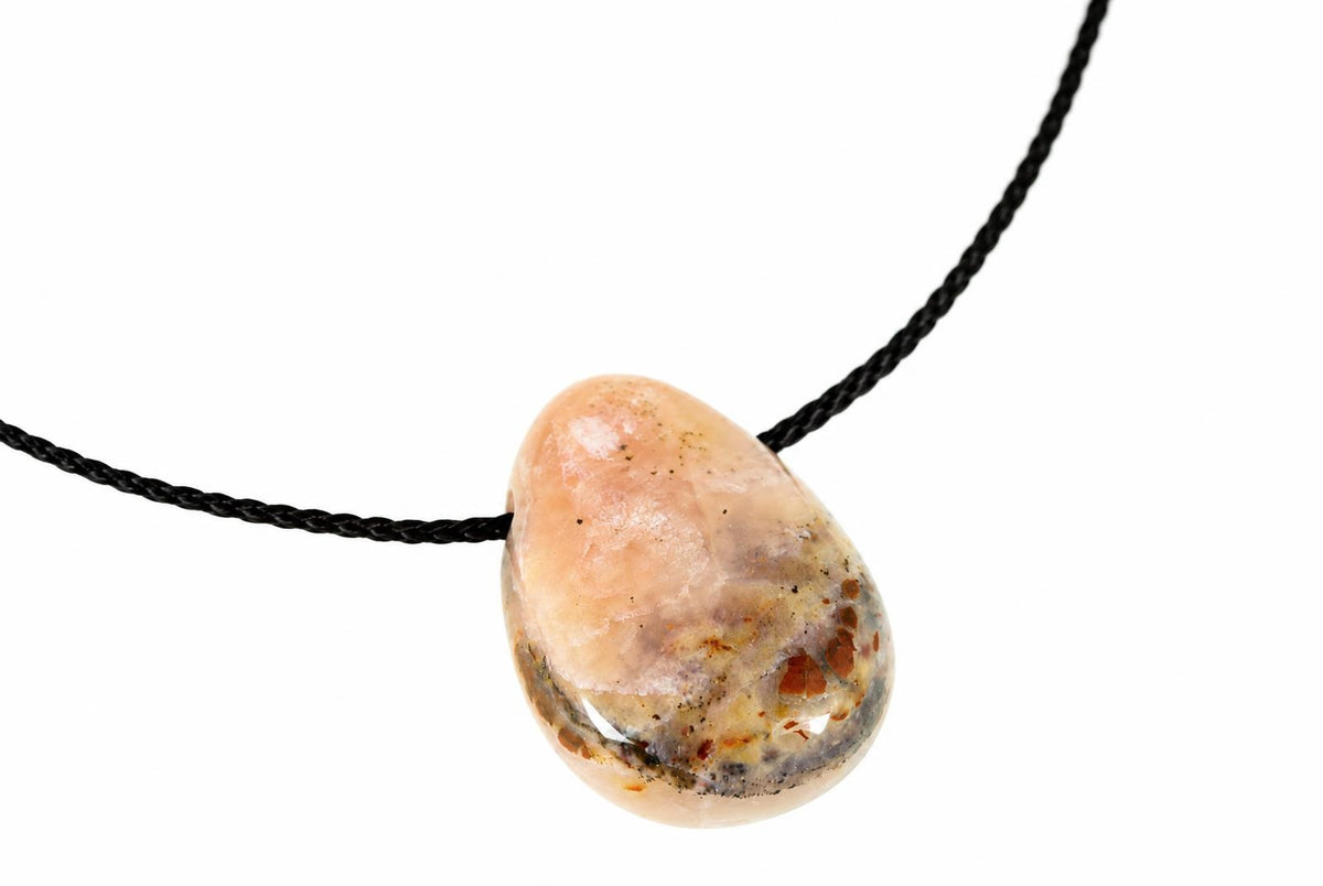 A teardrop-shaped pendant with a polished, peachy-pink stone speckled with brown and black inclusions on a dark cord against white.