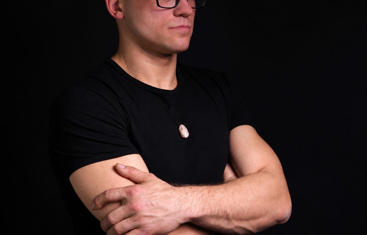A man in a black t-shirt wears a pendant with an oval, pale pink and white stone on a dark cord against a black background.