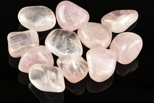 A collection of polished rose quartz stones in pale pink and clear tones, arranged on a dark reflective surface.
