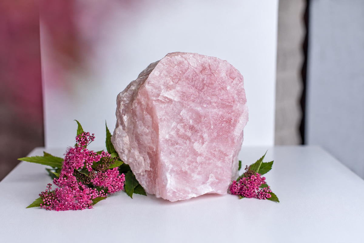 A large, rough pink crystal sits on a white surface with green leaves and small pink flowers nearby.