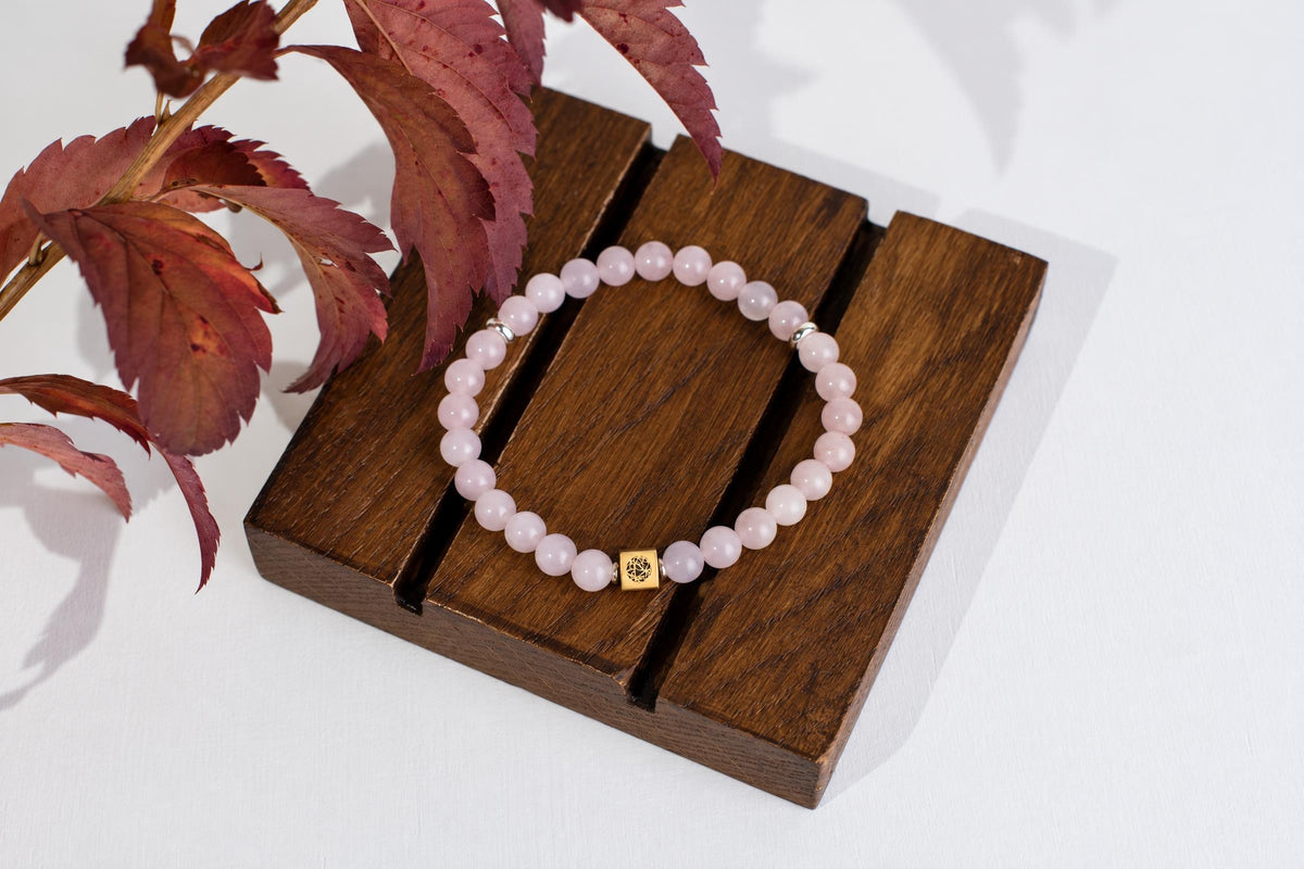 A bracelet with pale pink beads and a gold-toned square charm rests on a dark wooden surface, near reddish leaves.