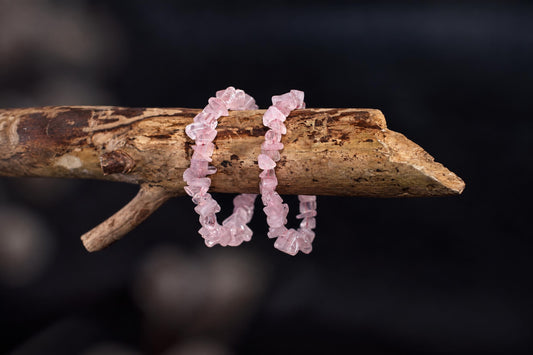 Two pink rose quartz chip bracelets rest on a rough wooden branch against a dark background.