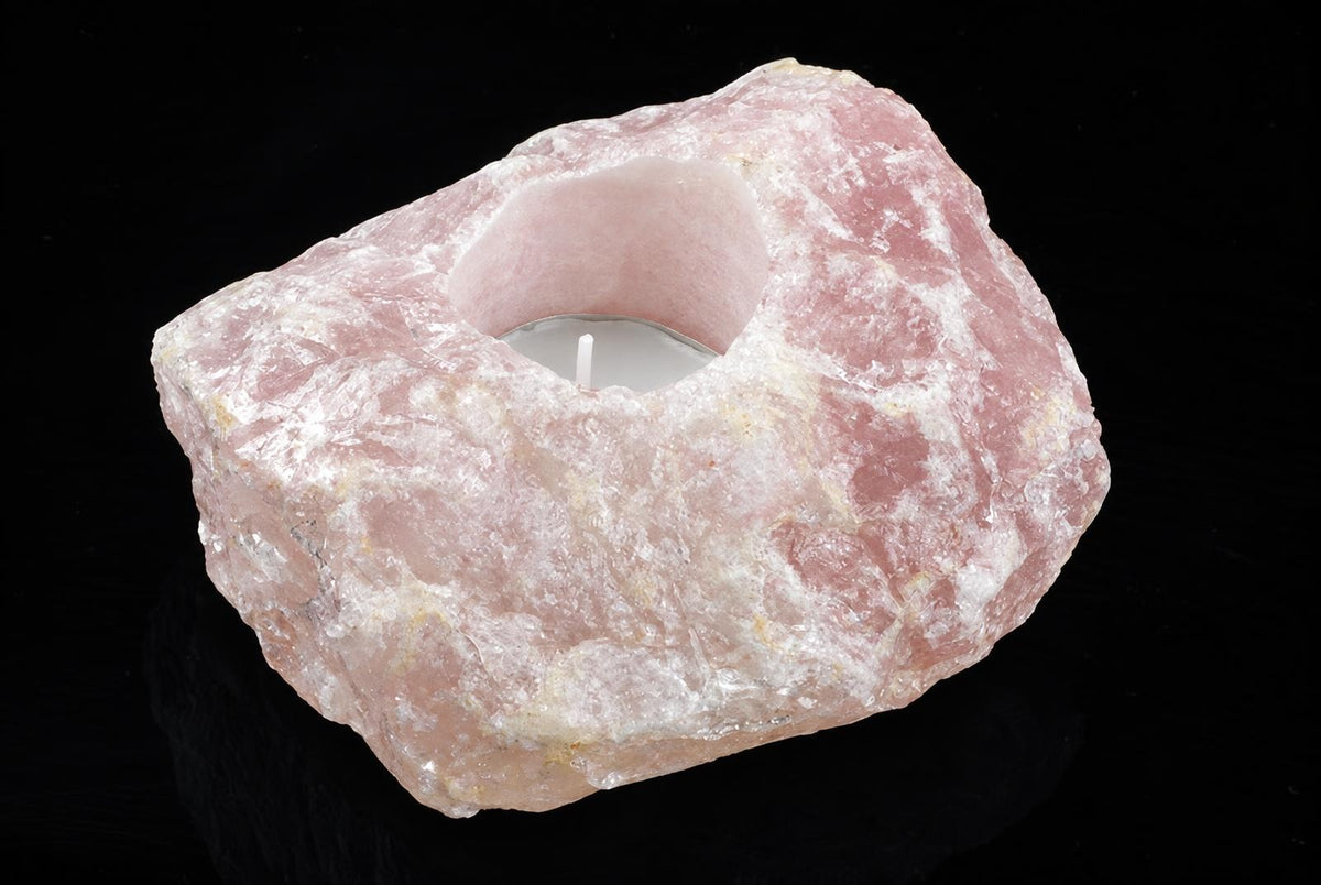 A rough, pink rose quartz stone with a carved cavity holding an unlit white candle on a dark surface.