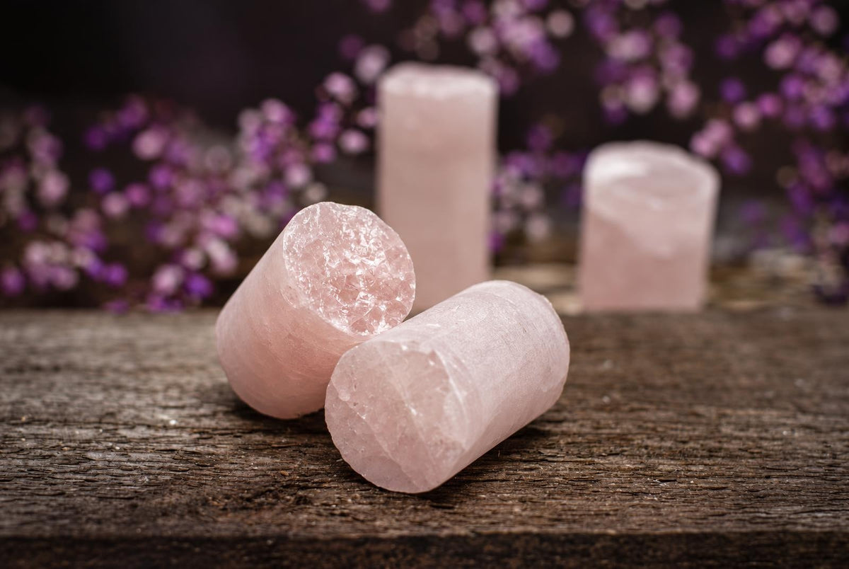 Three pink rose quartz drill cylinders rest on a dark wooden surface, with blurred purple flowers in the background.