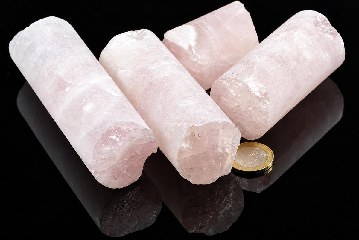 Pale pink rose quartz pieces with cylindrical forms and rough ends lie together on a glossy black surface beside a coin.