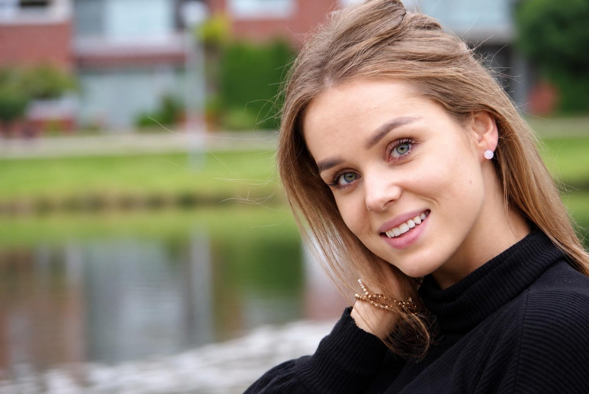 A smiling woman with light brown hair wears a black turtleneck and small white stud earrings against a blurred outdoor background.