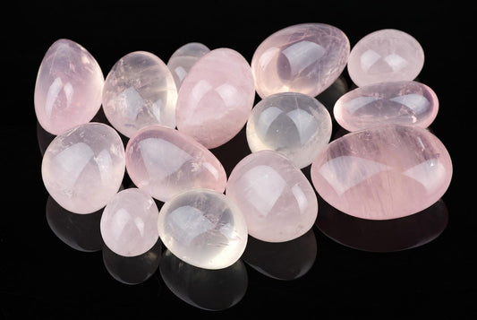A collection of polished rose quartz stones in shades of pink and clear, arranged on a dark reflective surface.