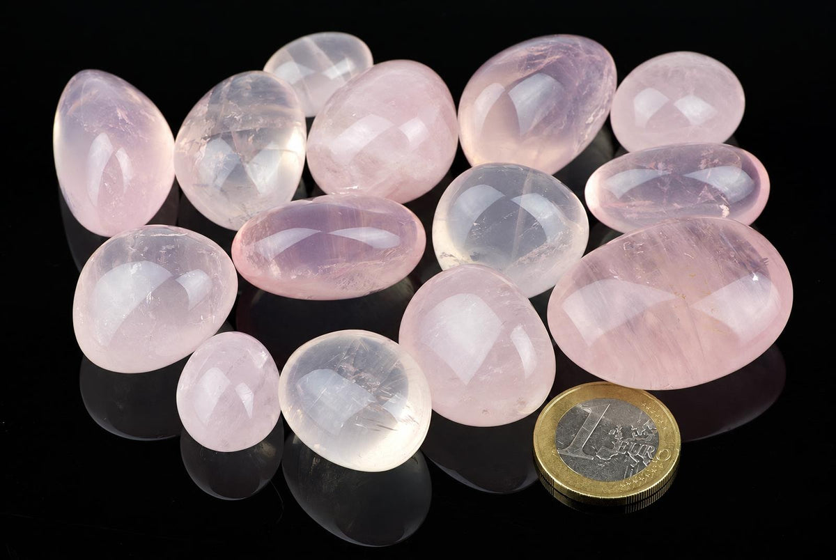 A collection of polished rose quartz stones in pink and clear shades, arranged on a dark reflective surface with a one euro coin for scale.