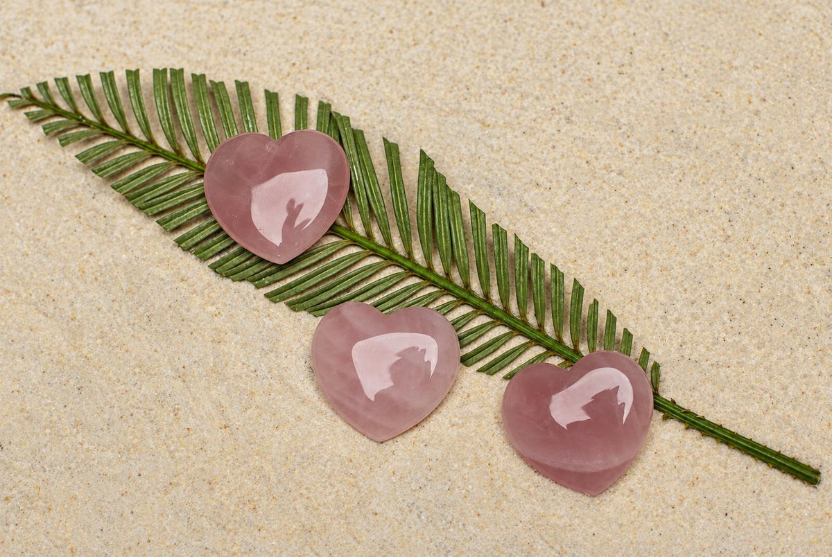 Three polished pink heart-shaped stones rest on a green leaf against a pale sandy surface.