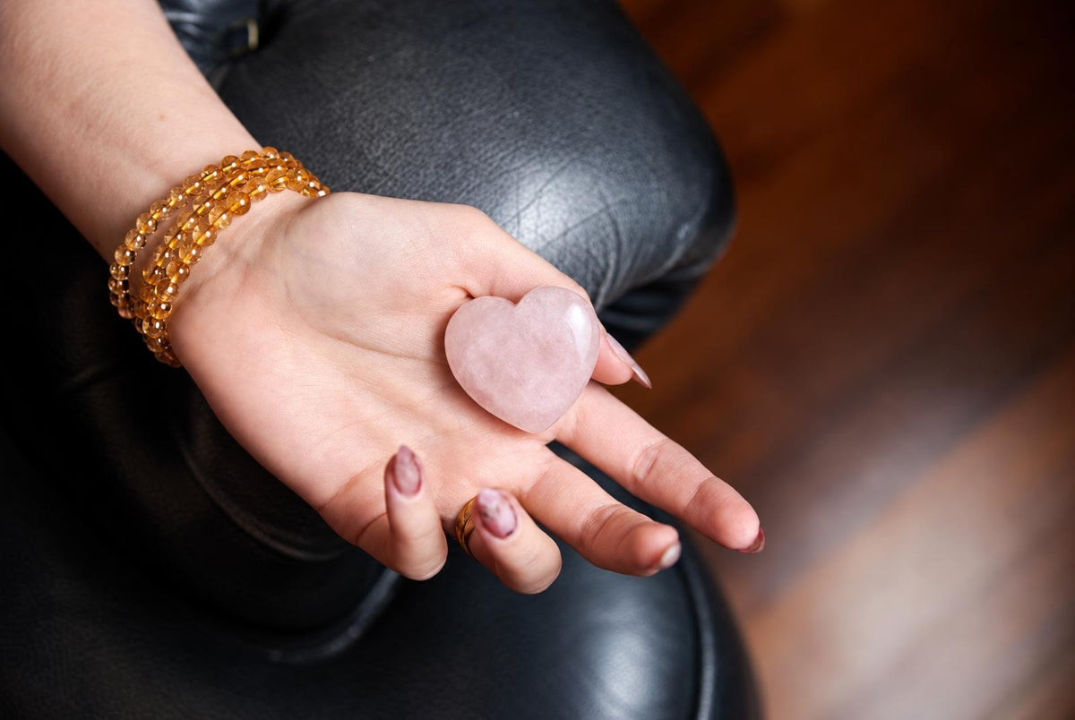 A hand holds a smooth, pink heart-shaped stone on a dark leather surface.