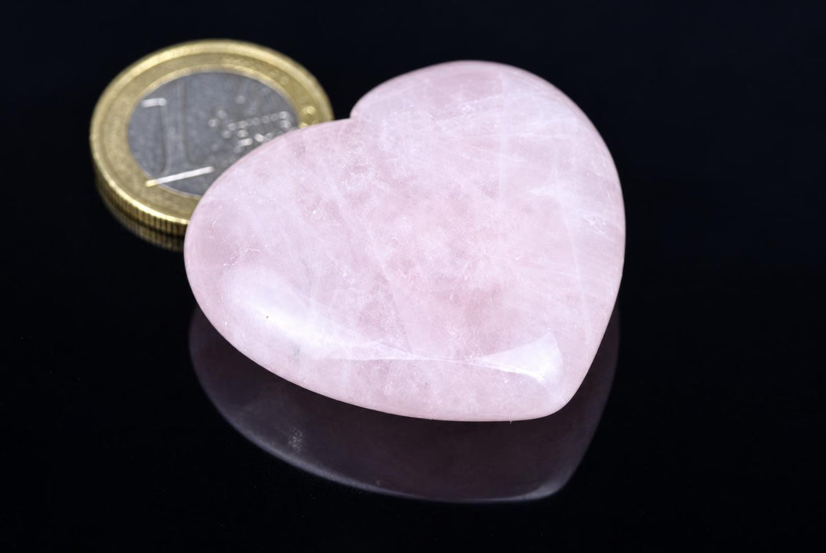 A polished pale pink rose quartz heart with cloudy white markings rests beside a coin on a glossy black surface.