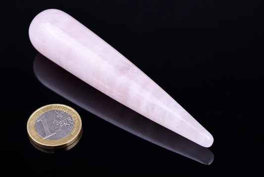 A pale pink, tapered massage stick rests on a dark surface next to a one-euro coin for scale.