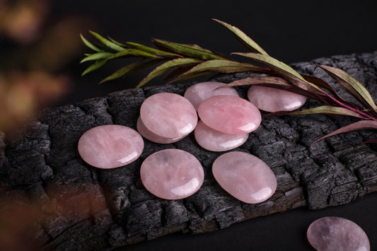 Several smooth, pink rose quartz palmstones are arranged on a dark wooden surface with greenery nearby.