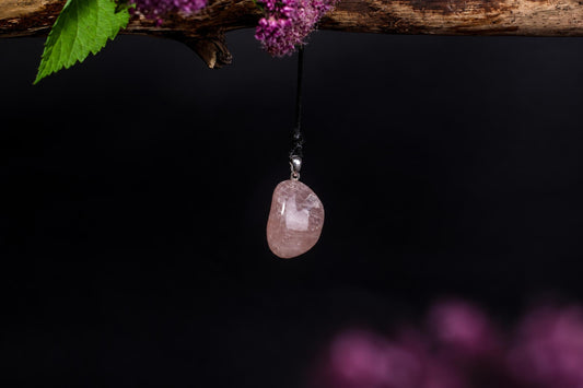 A pink rose quartz pendant hangs from a black cord against a dark background, with purple flowers and green leaves above.